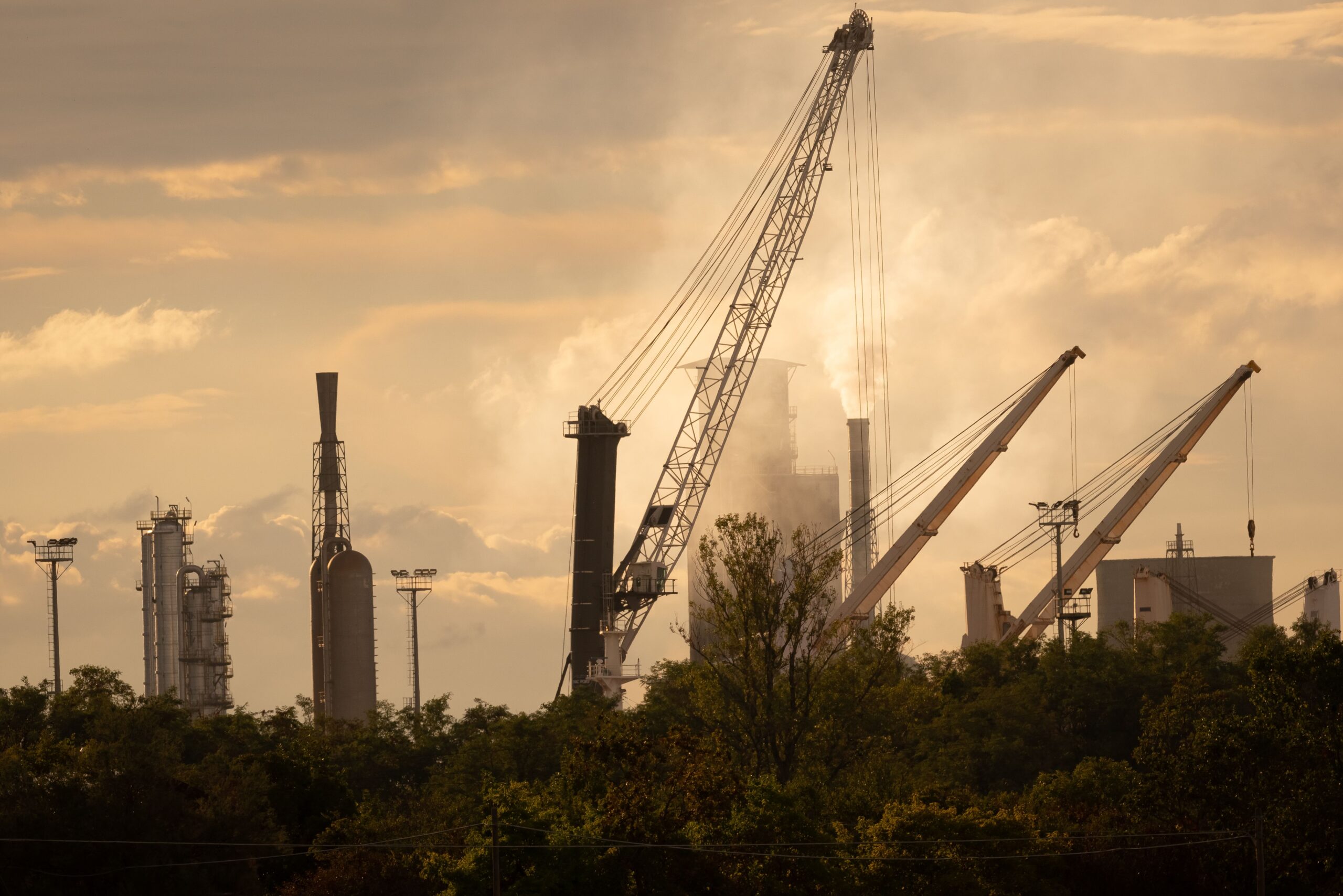 Industrial plant with tall stacks and cranes, steam rising behind trees under a hazy sky.