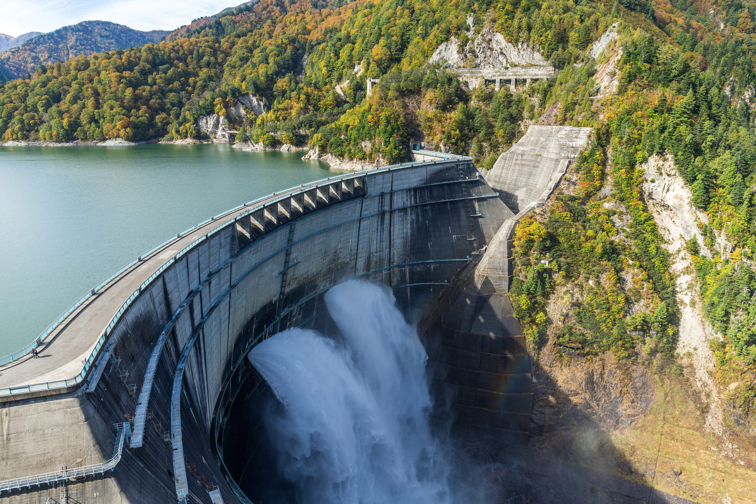 Curved concrete dam releasing water into a gorge beside a forested reservoir.